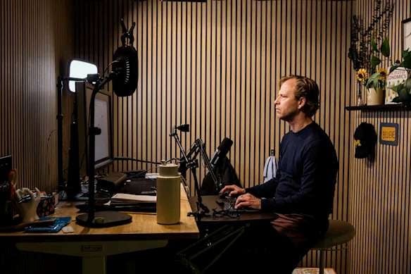 Justin Wolfers recording in his home studio in Ann Arbor, seated at a desk with microphone, screens and a timber-slat wall.